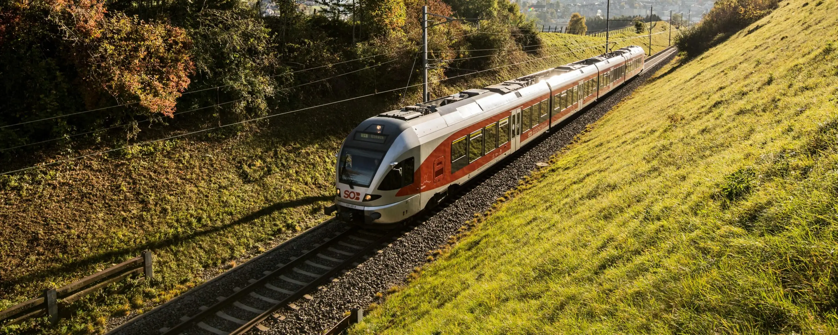 A red and silver regional train operated by the Swiss Südostbahn (SOB) travels through a green cutting on a sunny autumn’s day.