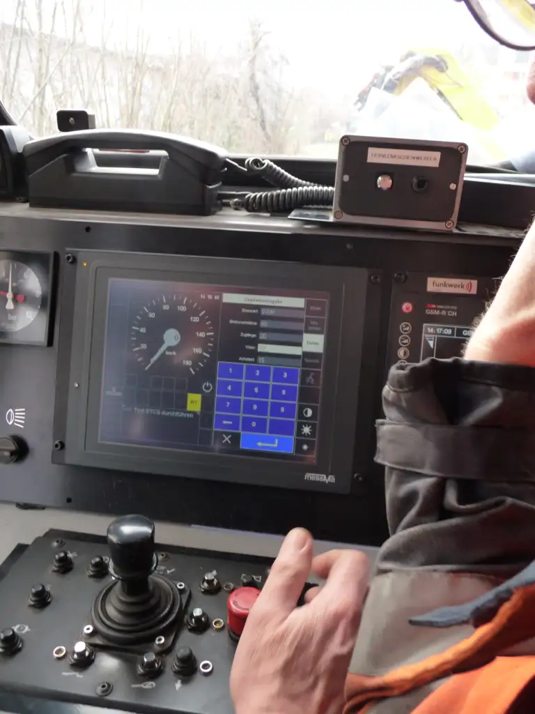Vue détaillée de l'intérieur d'un poste de conduite de locomotive moderne, montrant l'utilisation d'un écran tactile de contrôle et d'un joystick par le conducteur.
