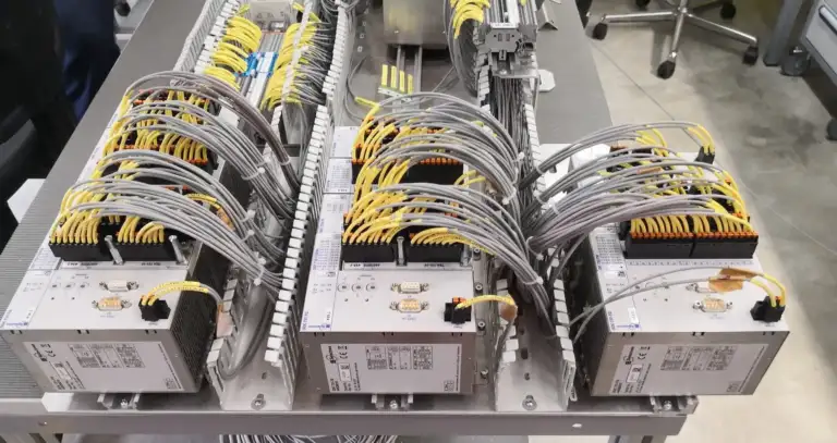 A close-up of an industrial switchgear and control unit featuring three Selectron enclosures, neatly arranged grey and yellow cable harnesses, terminal blocks and connectors on a metal table.