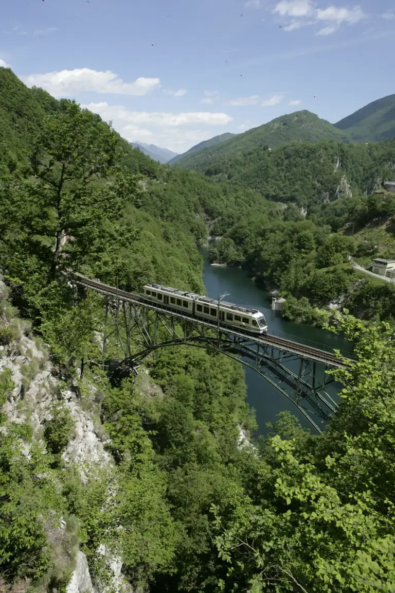 Ein FART-Regionalzug auf einer Stahlbrücke vor einem See in den Bergen