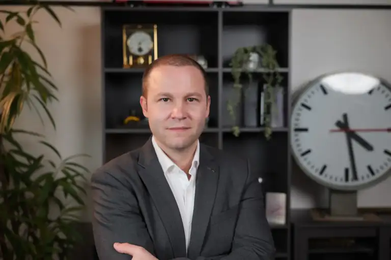 Portrait von Manuel Juon, der in einem Büro vor einem Bücherregal mit danebenstehender, grosser SBB-Uhr steht